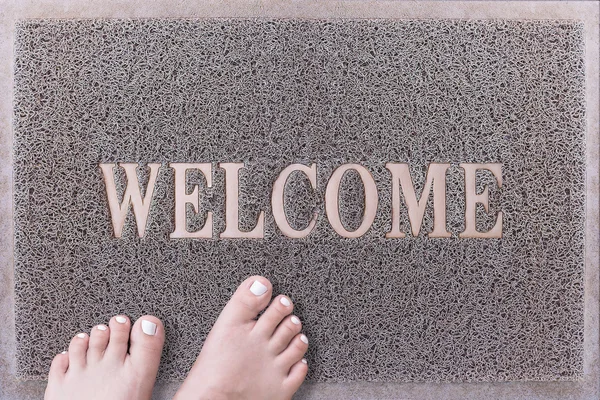 Welcome Door Mat With Female Feet. Friendly Grey Door Mat Closeup with Bare Woman Feet Standing. Welcome Carpet. Girl Feet with White Painted Toenails on Foot Scraper. — Stock Photo, Image