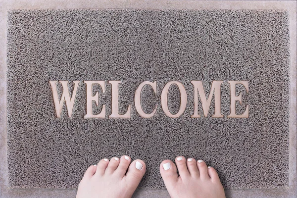 Welcome Door Mat With Female Feet. Friendly Grey Door Mat Closeup with Bare Woman Feet Standing. Welcome Carpet. Girl Feet with White Painted Toenails on Foot Scraper. — Stock Photo, Image