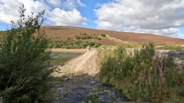 View of Hopes Reservoir Dam in the Lammermuir Hills of Scotland, showing the sturdy stone wall impounding the reservoir with surrounding grassland, distant hills, and clear sky overhead.