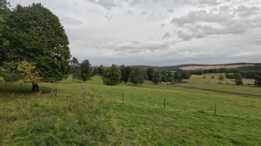 Footsteps along grassy field in the heart of Scottish Borders countryside, featuring open meadow with wildflowers, bordering dry stone walls, shallow burns, sheep trails, and expansive views over rounded hills and wooded valleys under clear skies.