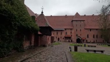 Polonya 'daki Malbork Kalesi' nin yağmurlu gündüz manzarası devasa tuğla duvarlar, Gotik kuleler ve bulutlu gökyüzünün altındaki Nogat Nehri 'ndeki hendekleri gösteriyor..
