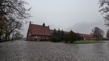 Polonya 'daki Malbork Kalesi' nin yağmurlu gündüz manzarası devasa tuğla duvarlar, Gotik kuleler ve bulutlu gökyüzünün altındaki Nogat Nehri 'ndeki hendekleri gösteriyor..