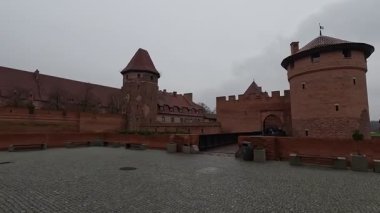 Polonya 'daki Malbork Kalesi' nin yağmurlu gündüz manzarası devasa tuğla duvarlar, Gotik kuleler ve bulutlu gökyüzünün altındaki Nogat Nehri 'ndeki hendekleri gösteriyor..