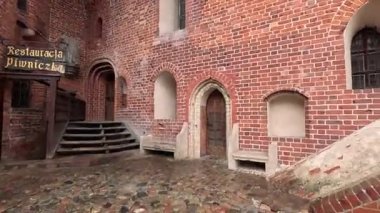 Polonya 'daki Malbork Kalesi' nin yağmurlu gündüz manzarası devasa tuğla duvarlar, Gotik kuleler ve bulutlu gökyüzünün altındaki Nogat Nehri 'ndeki hendekleri gösteriyor..