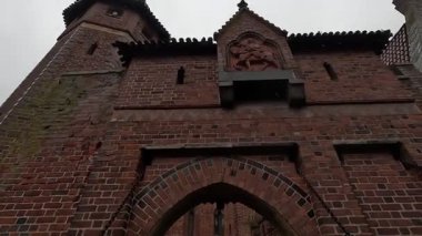 Polonya 'daki Malbork Kalesi' nin yağmurlu gündüz manzarası devasa tuğla duvarlar, Gotik kuleler ve bulutlu gökyüzünün altındaki Nogat Nehri 'ndeki hendekleri gösteriyor..