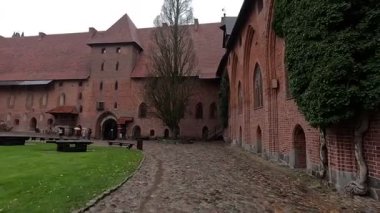 Polonya 'daki Malbork Kalesi' nin yağmurlu gündüz manzarası devasa tuğla duvarlar, Gotik kuleler ve bulutlu gökyüzünün altındaki Nogat Nehri 'ndeki hendekleri gösteriyor..
