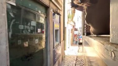 View from side window of historic yellow tram traveling along narrow streets in Lisbon, Portugal, passing old buildings, people and urban landscapes.