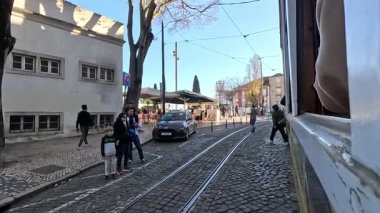 Lizbon, Portekiz 'de dar sokaklar boyunca seyahat eden tarihi sarı tramvayın yan penceresinden eski binaları, insanları ve kentsel manzaraları görüyoruz..