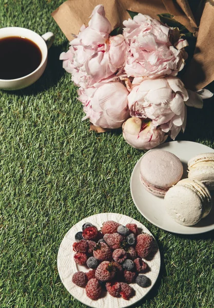 cup of coffee, berries, macaroons and peony flowers on green grass background