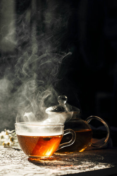 Warm cup of tea with glass teapot on dark background