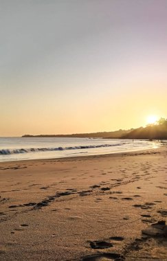 Sahilde gün batımı manzarası. Altın güneş, sahilin deniz ve kumlarını aydınlatıyor. Hafif dalgalar, bulutlu renkli gökyüzü ve sakin bir yaz ufku.