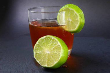 Buzlu bir bardakta servis edilen ferahlatıcı bir buzlu çayın yakın çekimi ve kenarında koyu arkaplanda izole edilmiş taze limon dilimi..