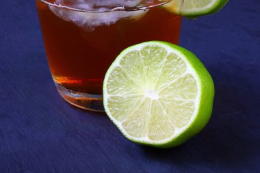 Buzlu bir bardakta servis edilen ferahlatıcı bir buzlu çayın yakın çekimi ve kenarında koyu arkaplanda izole edilmiş taze limon dilimi..