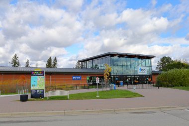 Winnipeg, Kanada 'daki Assiniboine Park Hayvanat Bahçesi' nin modern cam girişi. Parlak bulutlu gökyüzünün altındaki ana kapının yakınındaki patikalı sonbahar manzarası, bitkiler ve ziyaretçiler.