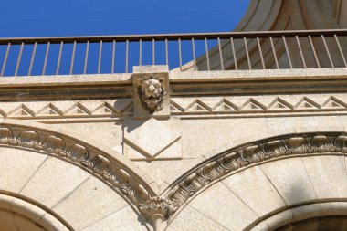 Kaya kemerleri, dekoratif oymalar ve berrak mavi gökyüzü altında klasik mimari unsurlar bulunan zarif tarihi bir binanın ön yüzü..