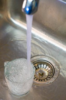 Temiz bardaklar, paslanmaz çelik lavaboda taze musluk suyuyla dolduruluyor. Temizliği, suyu, ev ortamını ve güvenli içme suyu konseptini simgeliyor..