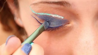 Bir makyözün elinin aşırı yakın plan çekimi bir kadının göz kapağına parlak mavi bir göz farı sürüyor.