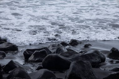 Okyanus dalgalarının ıslak kumda yumuşak siyah volkanik taşlarla kayalık bir kıyı şeridine ulaşmasını yakından görmek. Deniz köpüğü, hareket suyu ve karamsar atmosferli doğal sahil manzarası..