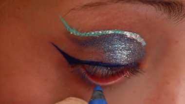 Bir makyaj sanatçısı su hattına mavi göz kalemi sürerken, bir kadının gözünün aşırı yakın çekimi.