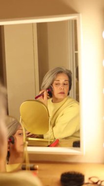 Gösterişli aynanın önünde gri saçlı, makyaj rutinini yapan yaşlı bir kadın.