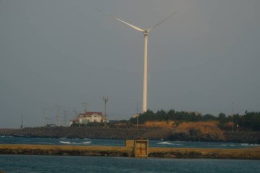 Jeju, Güney Kore - 11 Şubat 2026: Gün batımındaki Jeju cazibesi. Kıyıya yakın beton kuleler yel değirmeni