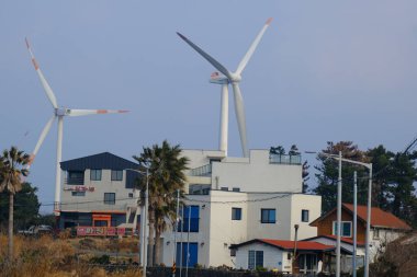 Jeju, Güney Kore - 11 Şubat 2026: Gün batımındaki Jeju cazibesi. Kıyıya yakın beton kuleler yel değirmeni