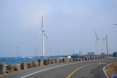 Jeju, Güney Kore - 11 Şubat 2026: Gün batımındaki Jeju cazibesi. Kıyıya yakın beton kuleler yel değirmeni