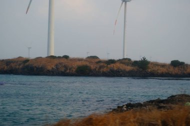 Jeju, Güney Kore - 11 Şubat 2026: Gün batımındaki Jeju cazibesi. Kıyıya yakın beton kuleler yel değirmeni