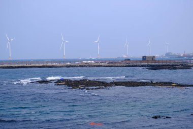 Jeju, Güney Kore - 11 Şubat 2026: Gün batımındaki Jeju cazibesi. Kıyıya yakın beton kuleler yel değirmeni