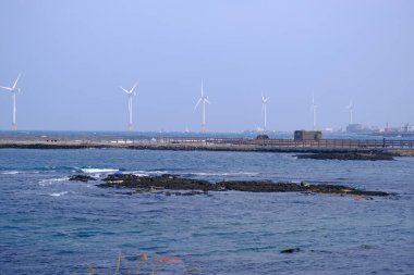 Jeju, Güney Kore - 11 Şubat 2026: Gün batımındaki Jeju cazibesi. Kıyıya yakın beton kuleler yel değirmeni