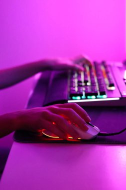 Pembe LED ışıklandırmada bilgisayar klavyesinde ve farede çalan ellerin yakın çekimi. Neon parıltısı ve siber estetiğe sahip modern oyun düzeneği.