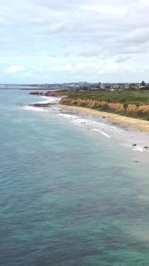 Uzun kumlu bir sahil sahili, okyanus dalgaları, uçurum kenarındaki bitki örtüsü ve bulutlu bir gökyüzünün altındaki uzak bir sahil kasabasının yüksek açılı görüntüsü..