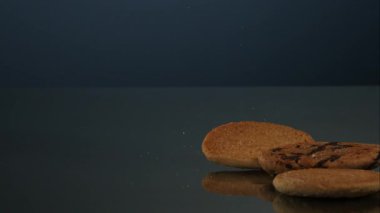 Bir sade altın kurabiye ve bir siyah çikolata parçalı kurabiye yukarıdan düşer ve koyu parlak yansıtıcı bir yüzeye sıçrar. Yüksek hızlı hareket yakalayan profesyonel bir stüdyoda diğer çerezlere katılır..