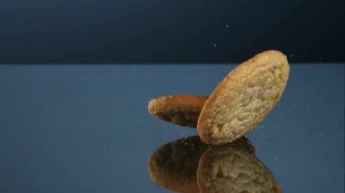 Yumuşak yuvarlak altın kurabiyeler yavaş çekimde uçan kırıntılar ve şeker parçacıklarıyla yansıtıcı gri bir stüdyo yüzeyinde düşüp zıplıyorlar.
