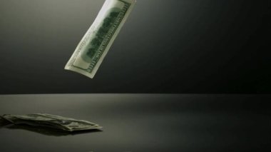 Finansal kaybı veya kazancı temsil eden gri bir arka plana karşı yavaş çekimde karanlık bir yansıtıcı yüzeye düşen yüz dolarlık banknot.
