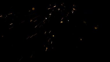 Parlak turuncu kıvılcımlar ve parlayan parçacıklar endüstriyel kaynak işleminden kalma siyah bir zemine karşı yavaş çekimde havada uçuyor.