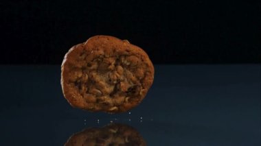Tek parça çikolatalı yulaf ezmeli kurabiye koyu renkli yansıtıcı bir yüzeye düşüp kırıntıları aşırı yavaş çekim stüdyo çekimine serpiştiriyor.
