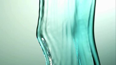 Dikey turkuaz renkli su akışı, parlak beyaz ve yeşil arkaplana karşı yavaş çekimde birçok küçük damlacığa dökülüp kırılıyor.