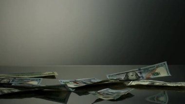Diğer banknotların yavaş çekimde yattığı gri bir yüzeye havadan büyük miktarda 100 dolarlık banknotlar düşüyor.
