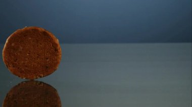 Tek bir yulaflı kurabiye düzgün mavi bir zemin üzerinde yansıtıcı karanlık bir yüzey üzerinde yavaşça ve sabit dairesel bir hareketle ekran için yuvarlanıyor.