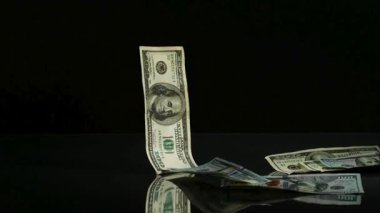 Yüz dolarlık banknotlar tek tek siyah yansıtıcı zemine dökülüyor. Stüdyo ortamında dağınık bir para yığını oluşturuyor.