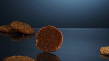 Tek bir yuvarlak yulaflı kuru üzümlü kurabiye, arka planda diğer kurabiyelerle birlikte koyu mavi yansıtıcı yüzeyde ağır çekimde döner ve döner.