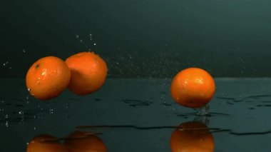 Çok sayıda taze turuncu mandalina düşer ve ıslak bir yansıtıcı yüzeyde zıplar. Su sıçrar ve damlalar yavaş çekimde koyu bir zemine çarpar.
