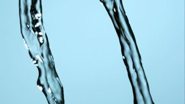 Kristal berrak sıvı su, dans eden ışık yansımaları olan parlak mavi bir arka plana karşı dinamik bir şekilde yüksek hızda yavaş çekimde sıçrar ve akar.