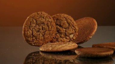 Fırınlanmış taze yulaf ezmesi ve çikolata parçalı kurabiyeler yavaş çekimde karanlık bir yansıtıcı yüzeye dökülüp duruyor.