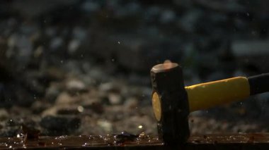 Ağır balyoz, kahverengi cam bir bira şişesini kırıyor. Tahta bir yüzeyde su sıçratıyor ve cam parçaları yavaş çekimde uçuyor.