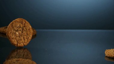 Lezzetli yuvarlak yulaf ezmeli kuru üzümlü kurabiyeler, yemek reklamları için yavaş çekimde yumuşak mavi bir arka plana karşı koyu bir yansıtıcı yüzeyde yuvarlanıp hareket ediyorlar.