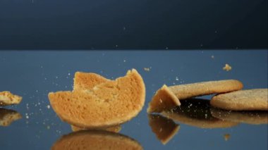 Tek bir altın yuvarlak kurabiye koyu bir yansıtıcı yüzeyde düşüp kırılıyor ve kırıntılar mavi bir stüdyonun arka planına saçılıyor.