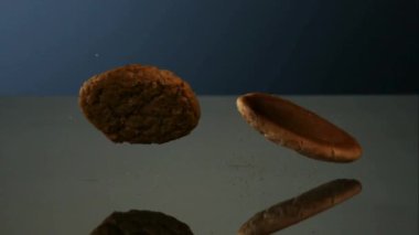Taze pişmiş yulaf ezmesi ve çikolata parçalı yuvarlak kurabiyeler yumuşak mavi ve gri stüdyo ışığı ile yansıtıcı bir ayna yüzeyine düşüp zıplıyorlar.