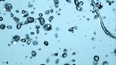 Temiz su damlacıklarının ve baloncukların yüksek hızda makro görüntüsü açık mavi bir arkaplana doğru düşerek canlandırıcı bir soyut doku ve hareket yaratır.